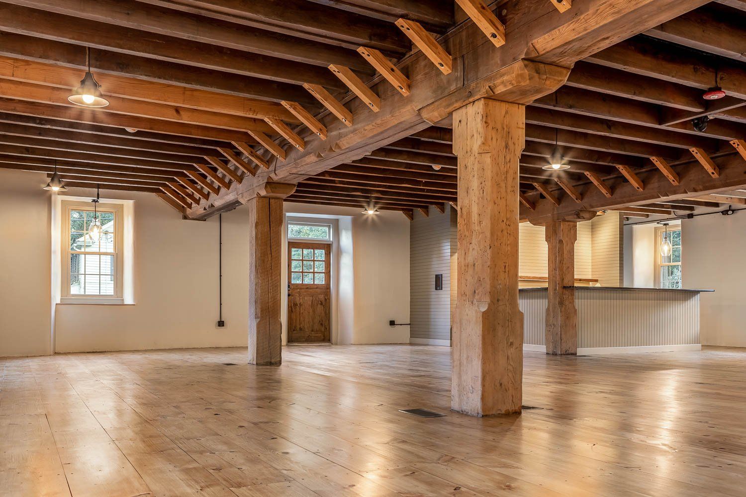 Spacious interior with exposed wooden beams, support pillars, and hardwood floors. Door and windows provide natural light.