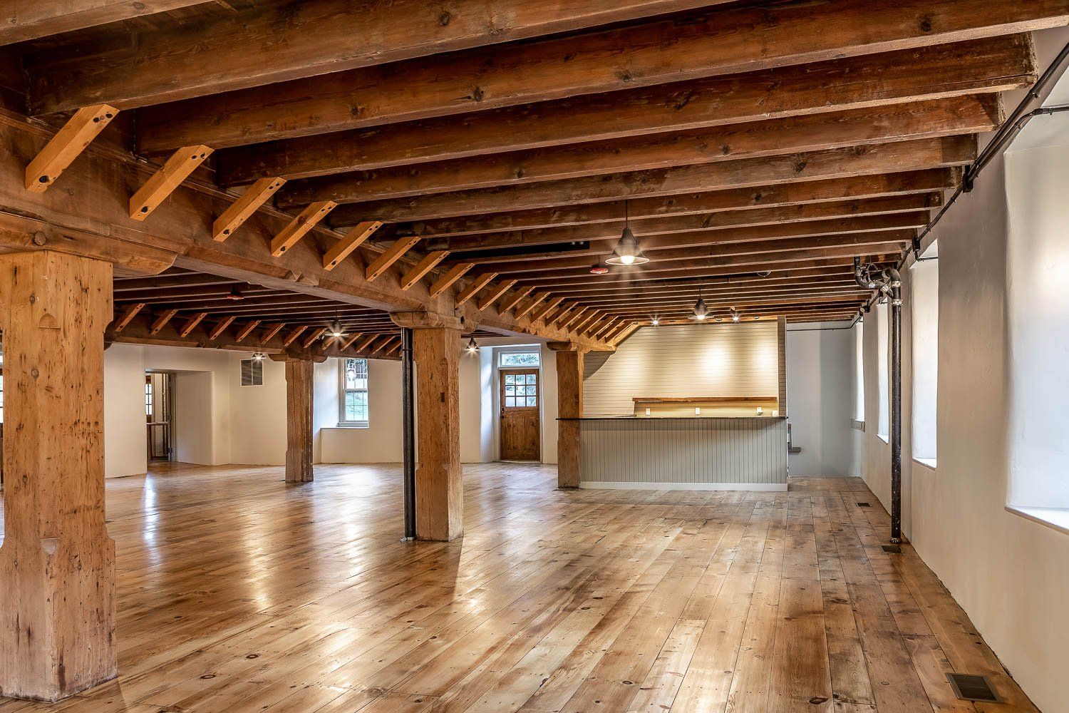 Spacious room with wooden floors, exposed beams, and supporting columns. White walls and natural light.