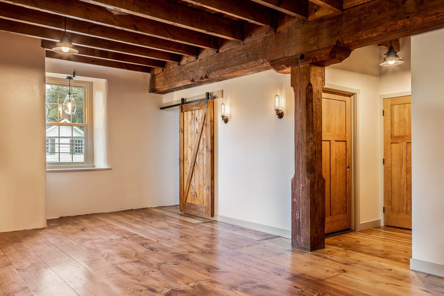 Interior of a room with wood beams, plank floors, and barn doors. Features a window, doors, and sconce lighting.