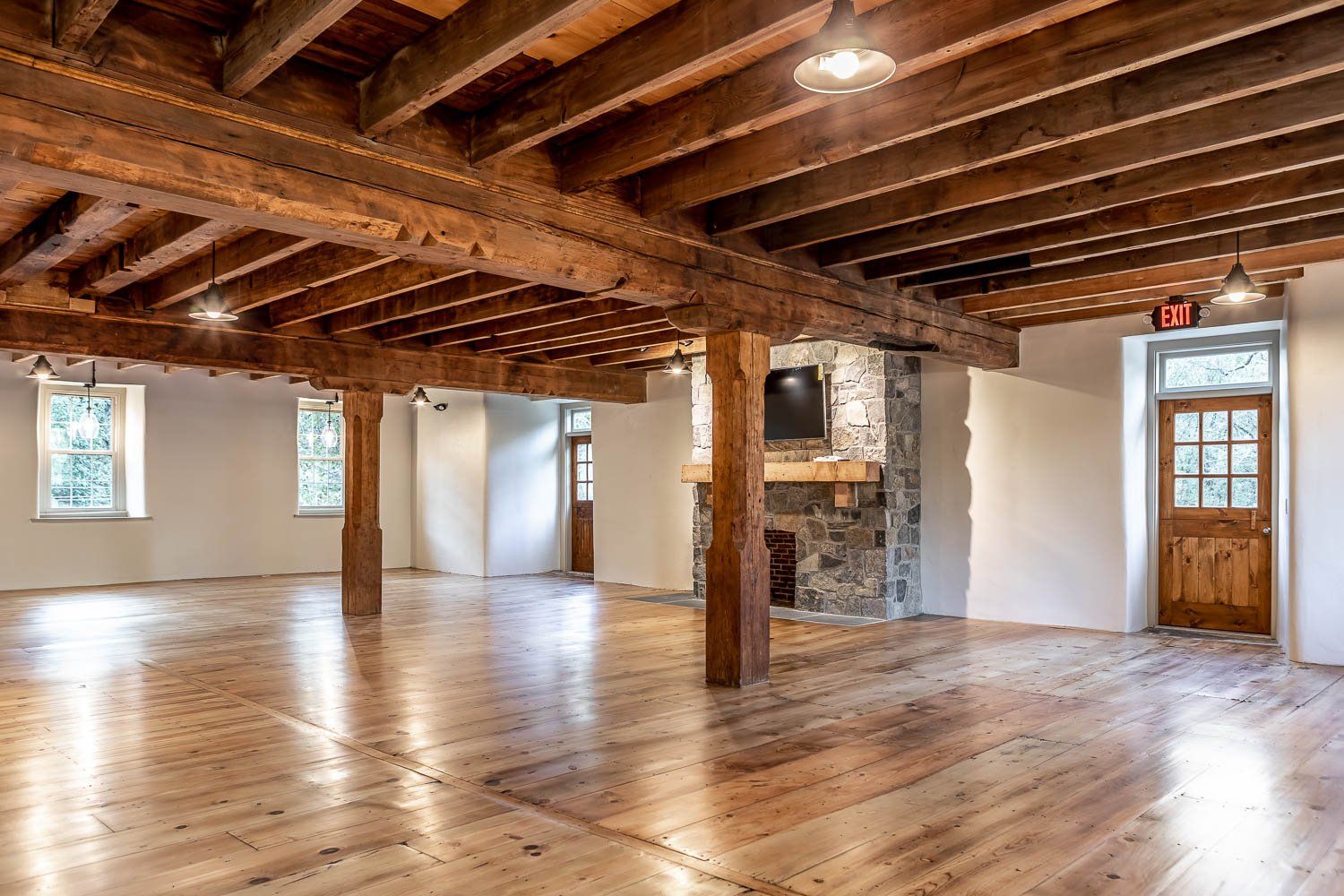Spacious room with wood beams, hardwood floor, stone fireplace, and windows.