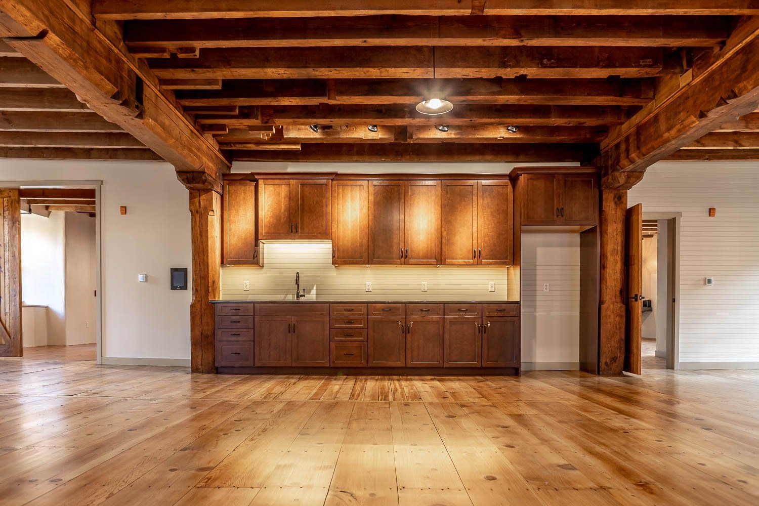 Wooden kitchen cabinets in a spacious room with exposed wooden beams, hardwood floors, and empty doorways.