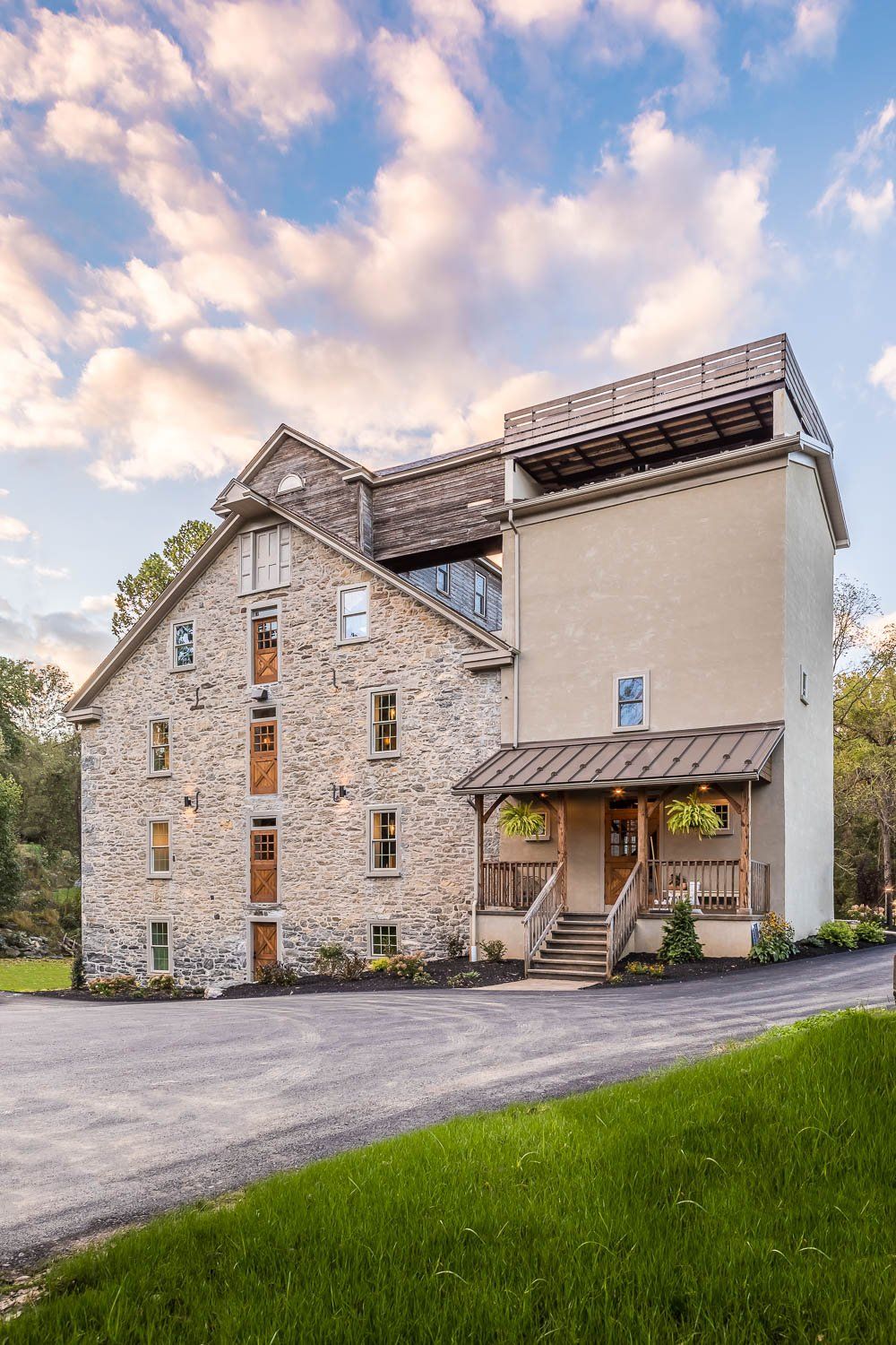Stone and tan building with wooden porch, gravel drive, and green grass against a cloudy sky.