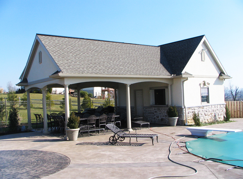 Poolside cabana with an open-air dining area, seating, and lounge chair. The building has a gray roof and stone accents.