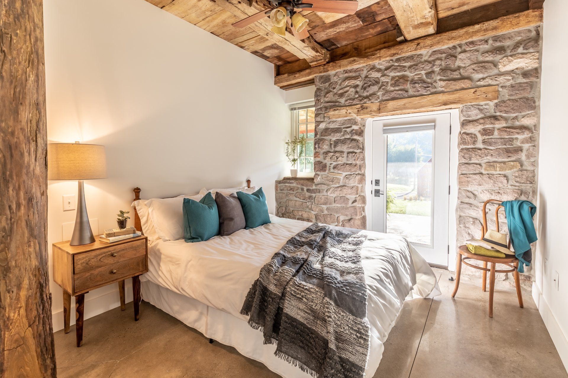 Bedroom with stone wall, wooden beams, bed with pillows and blanket, nightstand, and chair.