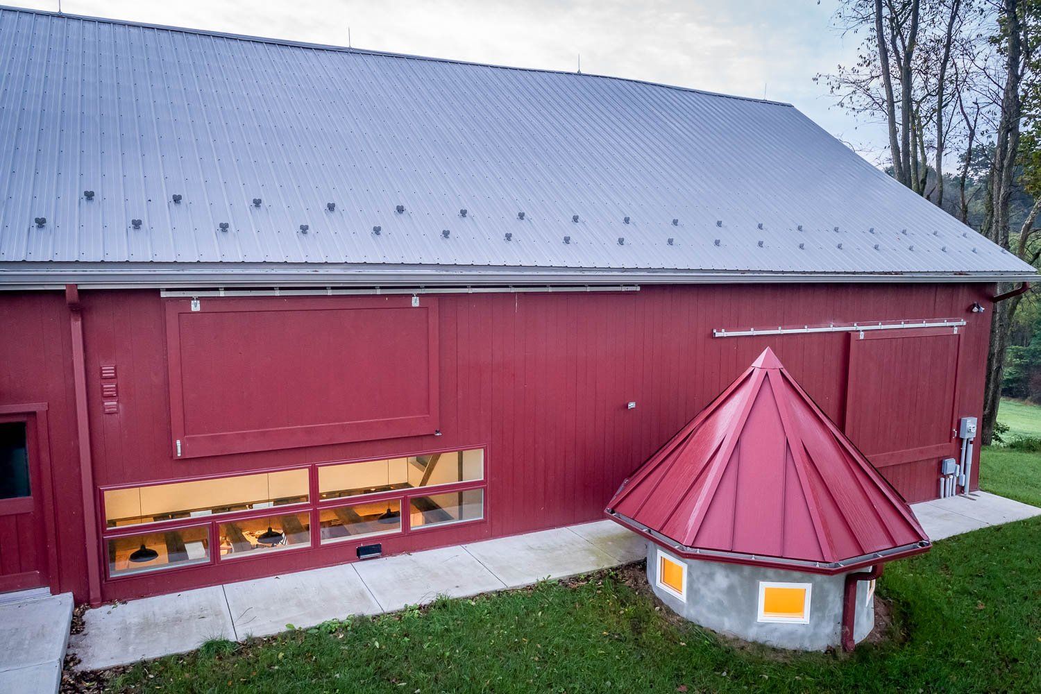 Red barn with a metal roof and small attached building with a cone-shaped roof on a grassy lawn.