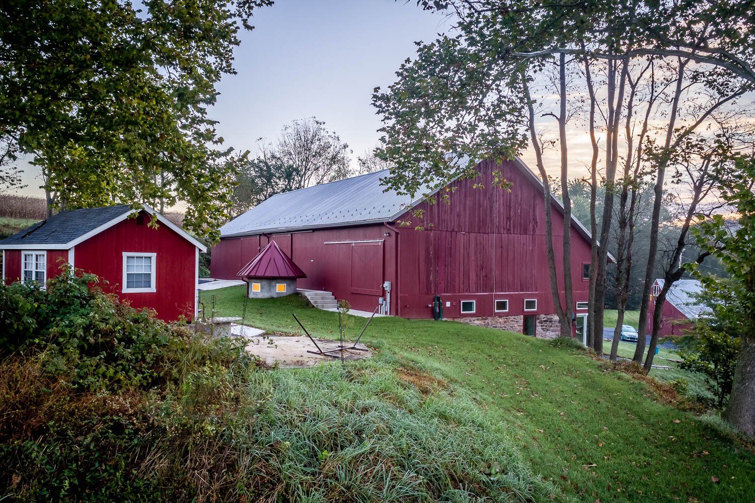 Red barn and small red shed on a grassy hill, trees in the background, overcast sky.