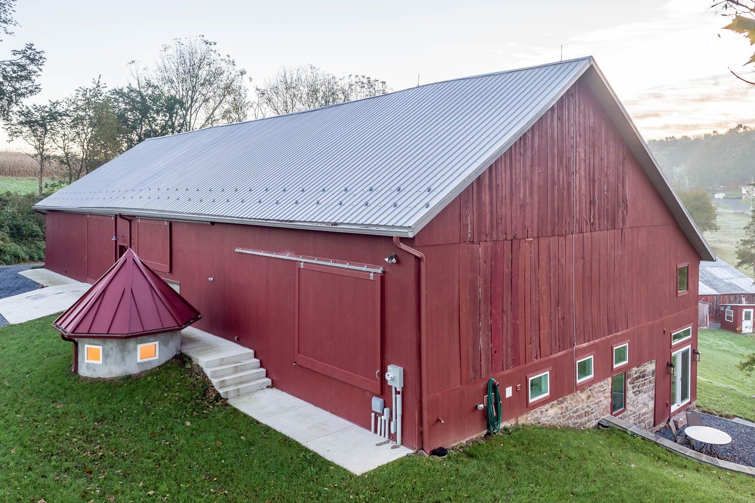 Red barn with corrugated metal roof, steps leading to a small, turret-like structure. Green lawn, trees in background.
