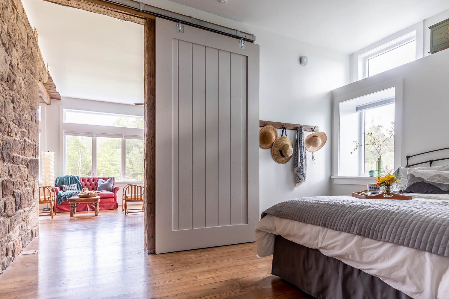 Bedroom with barn door leading to living room, bed with gray bedding, hats on wall.