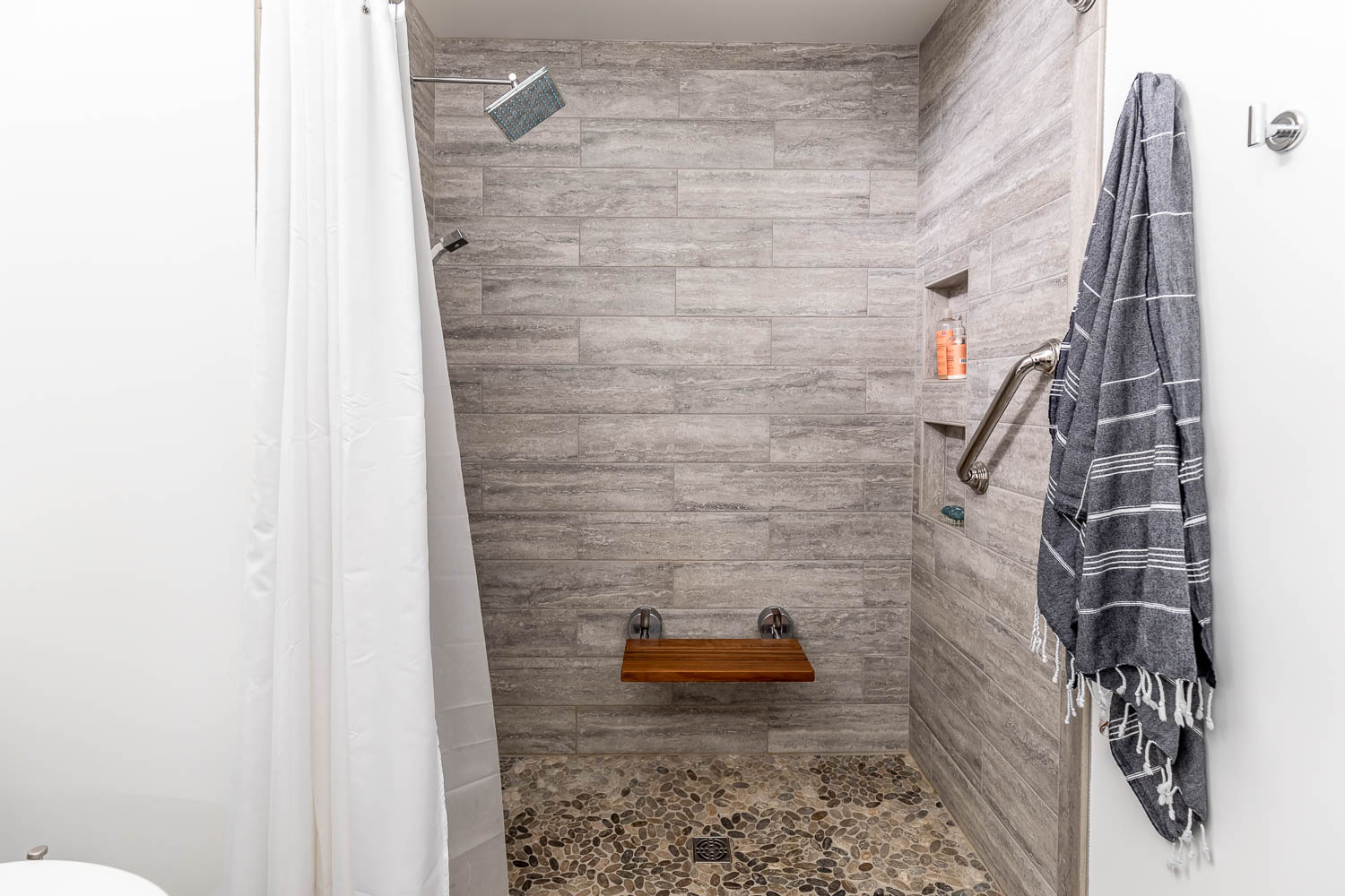 Shower stall with stone tile walls, wooden seat, and towel hanging on the right.