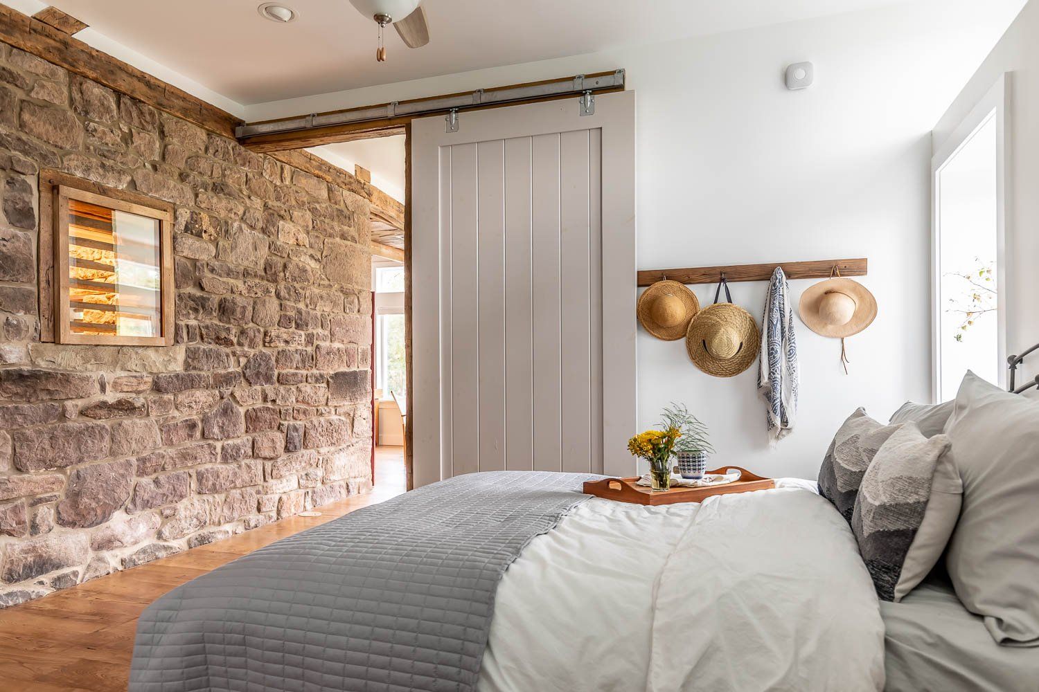 Bedroom with stone wall and sliding door, bed with gray comforter, hats hanging on wall.