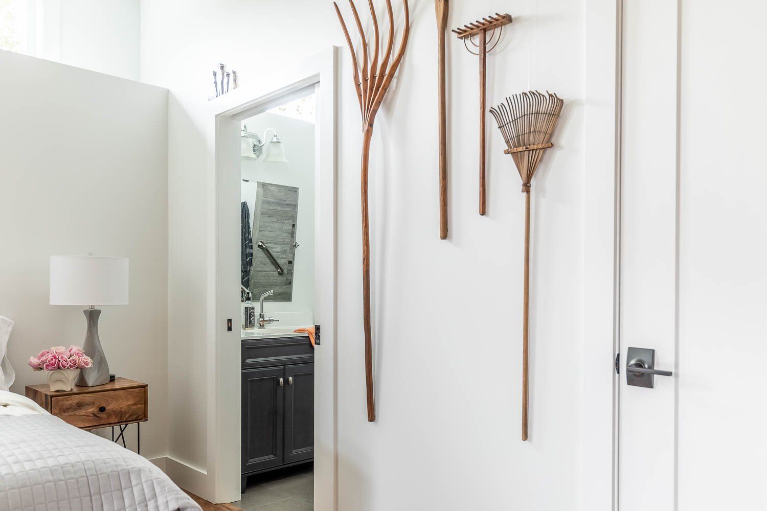 Bedroom with white walls, three rustic tools on display, a nightstand, and a glimpse of a bathroom.