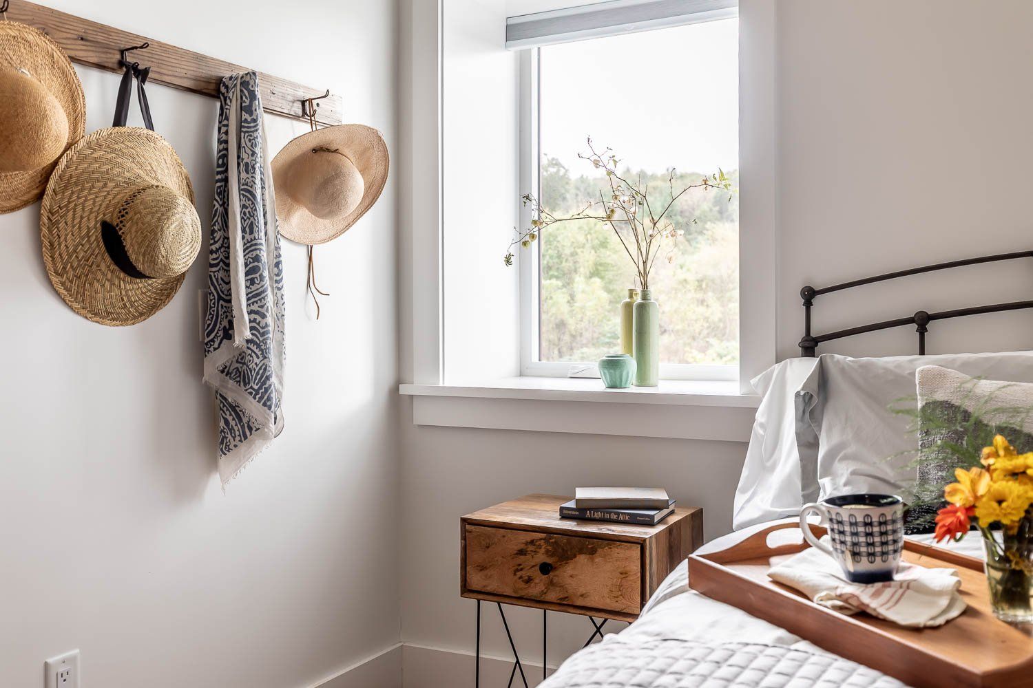 Bedroom with bed, nightstand, window, hats hanging on a wood rack, and a tray with items.