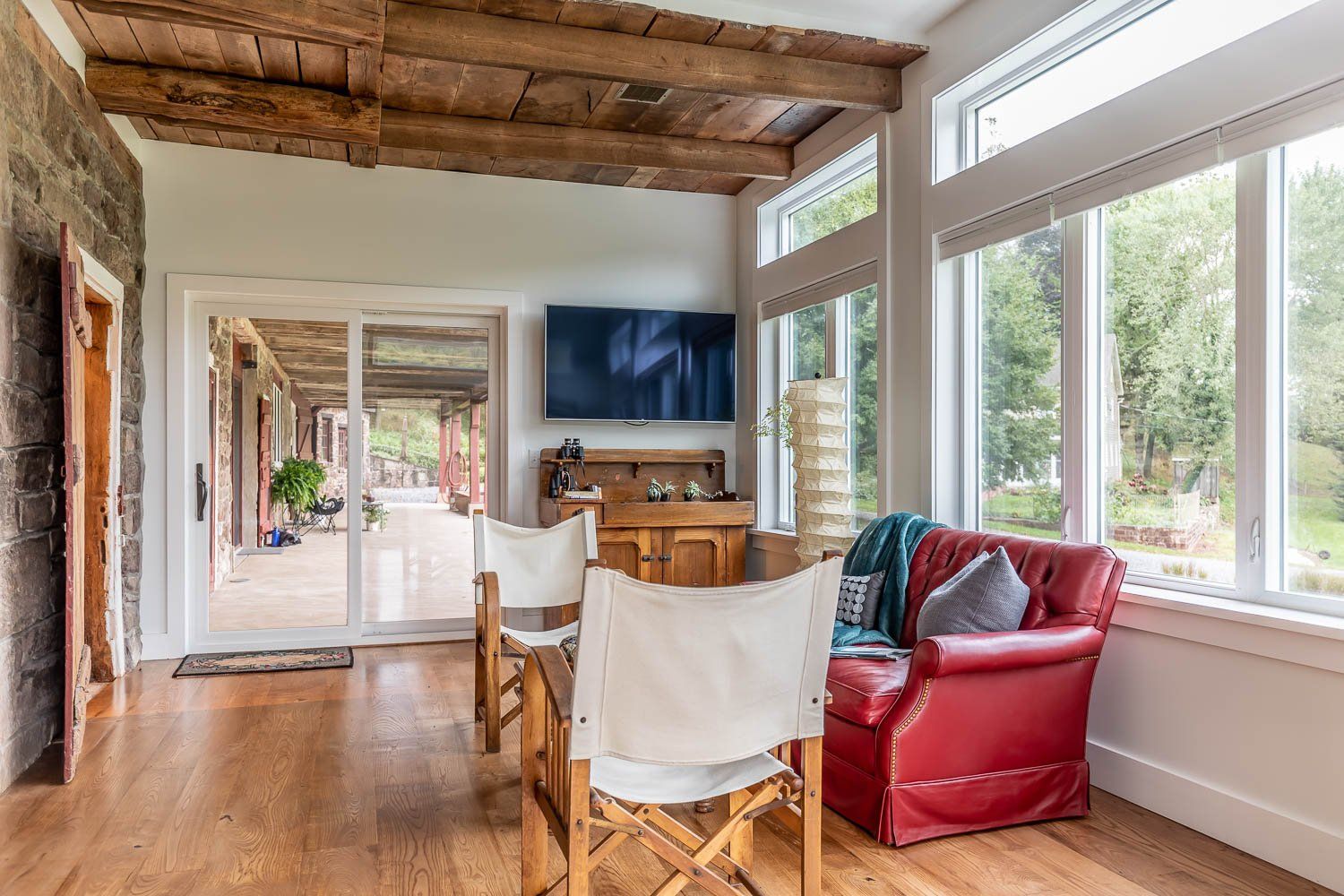 Sunroom with wood beams, red leather chair, table, and large windows.
