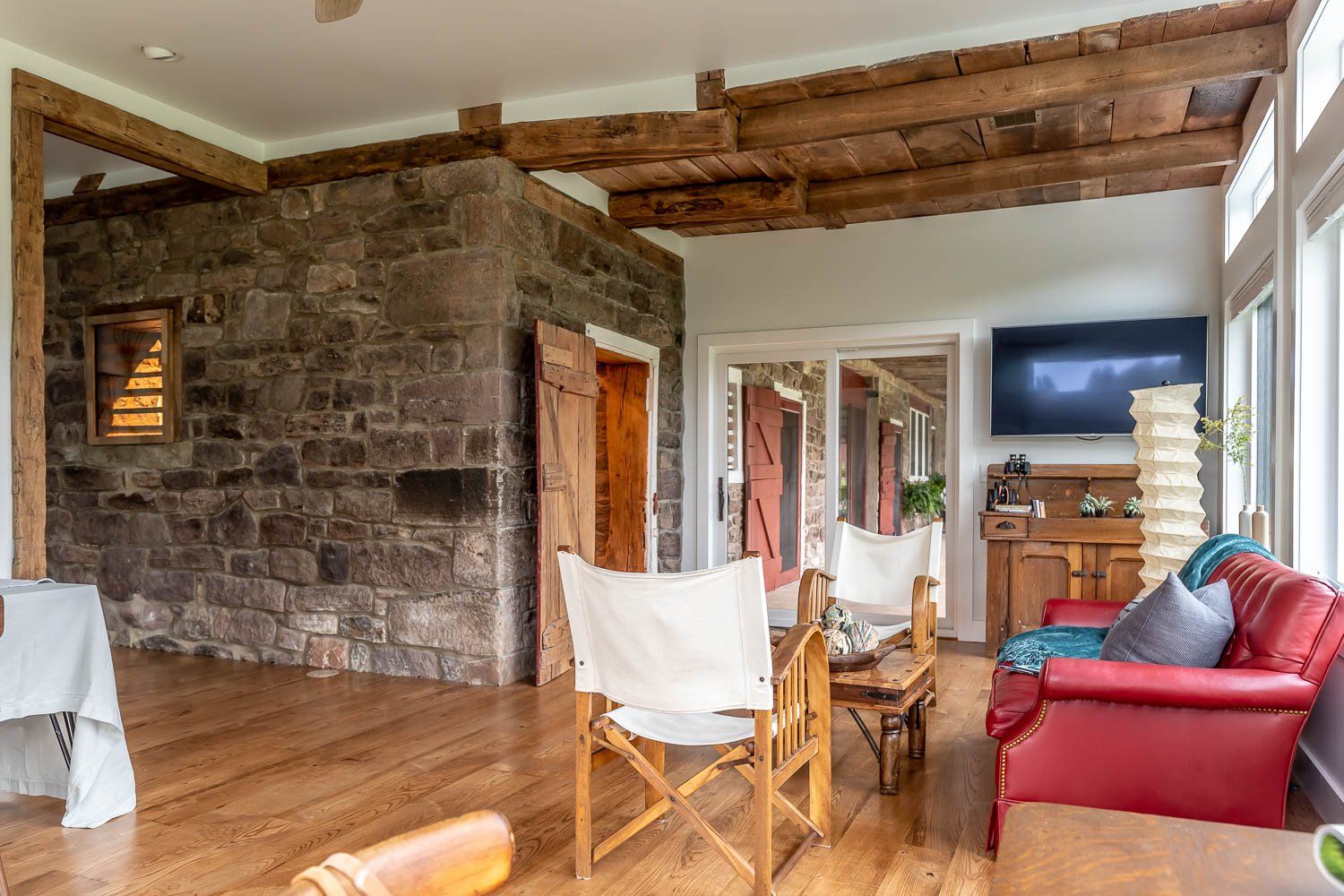 Sunroom with exposed stone wall, wood beams, red leather sofa, wood chairs, and TV.