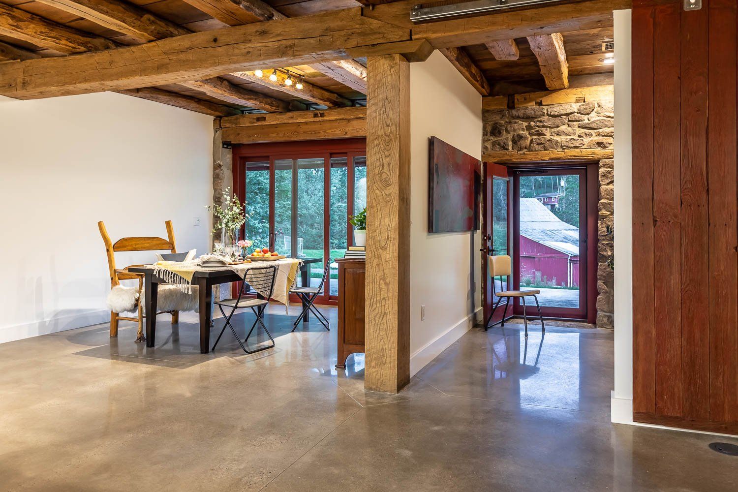 Open-concept dining area with exposed wood beams, polished concrete floor, red door, stone wall, and dining table set.