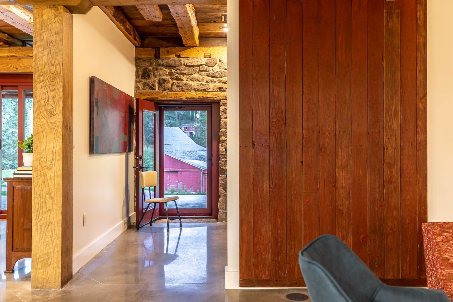 Interior view: hallway with stone wall, wood paneling, open door to a red building, concrete floor, and art.