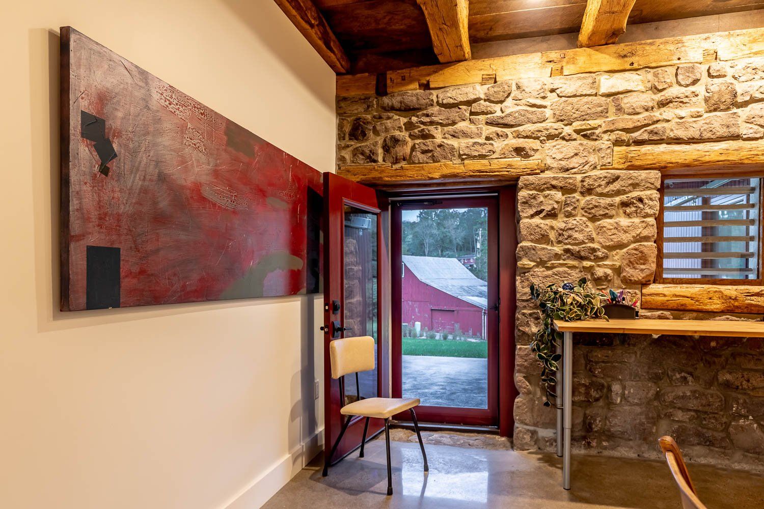 Interior with red door open to a barn, abstract art, stone wall, wood beams, and a chair.