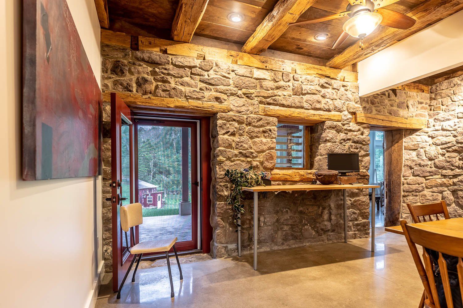 Stone-walled room with wooden beams and a doorway leading to a porch. A chair sits by the open door.