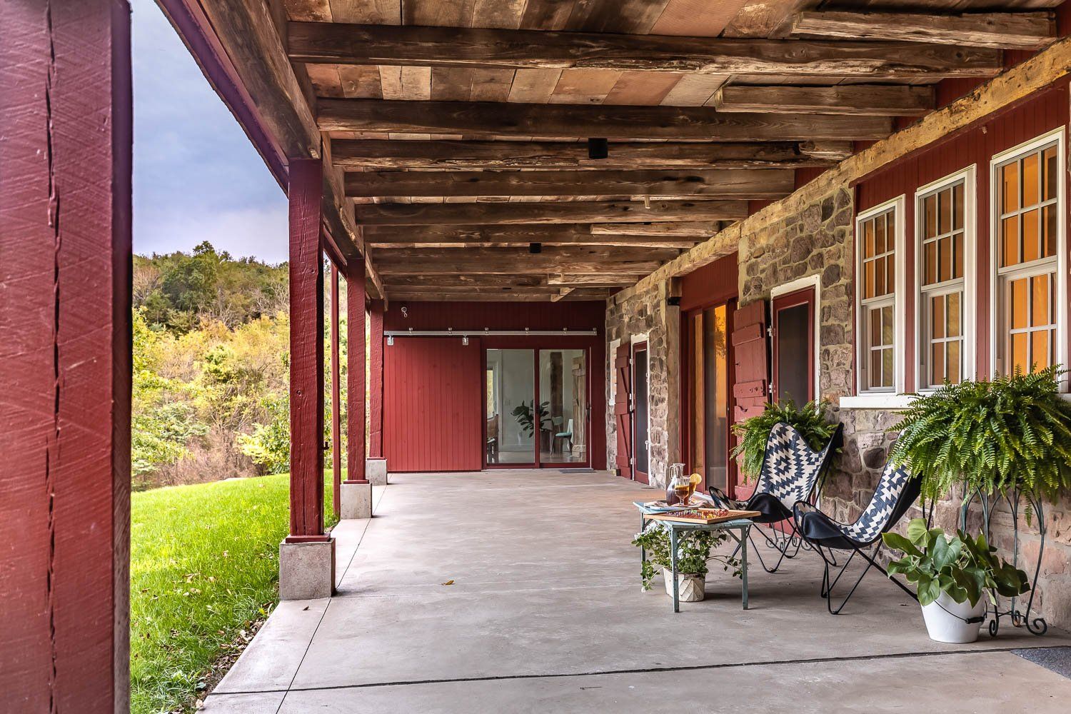 Covered porch of a stone building with red trim. Two chairs sit next to table. Greenery and a grassy lawn are visible.