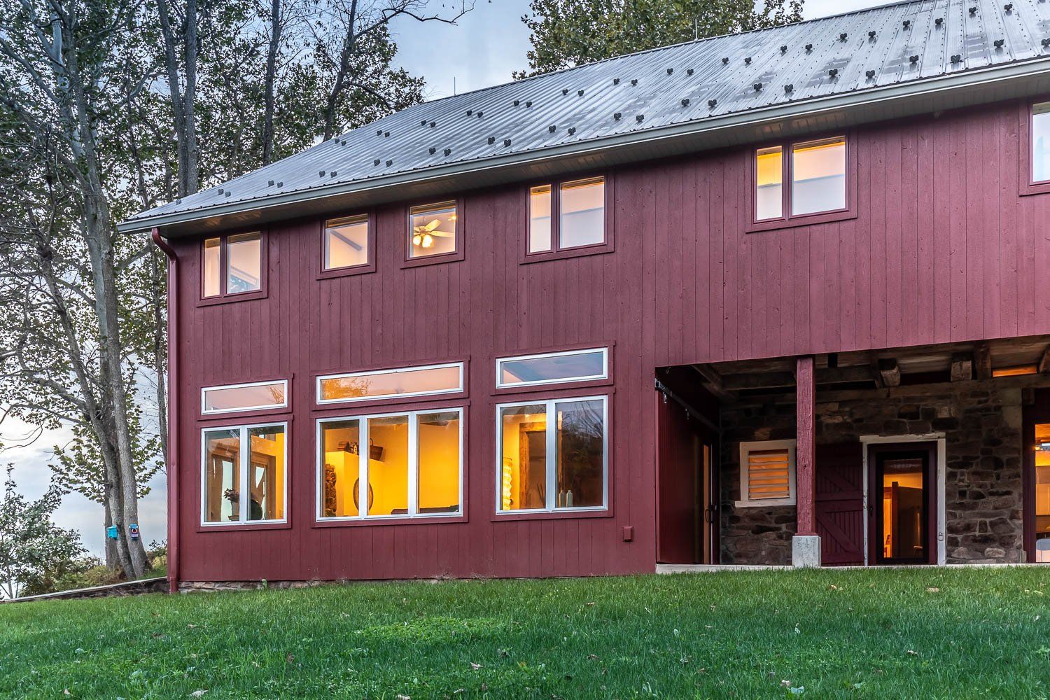 Two-story red building with multiple windows, metal roof, and stone base. Green grass in front.