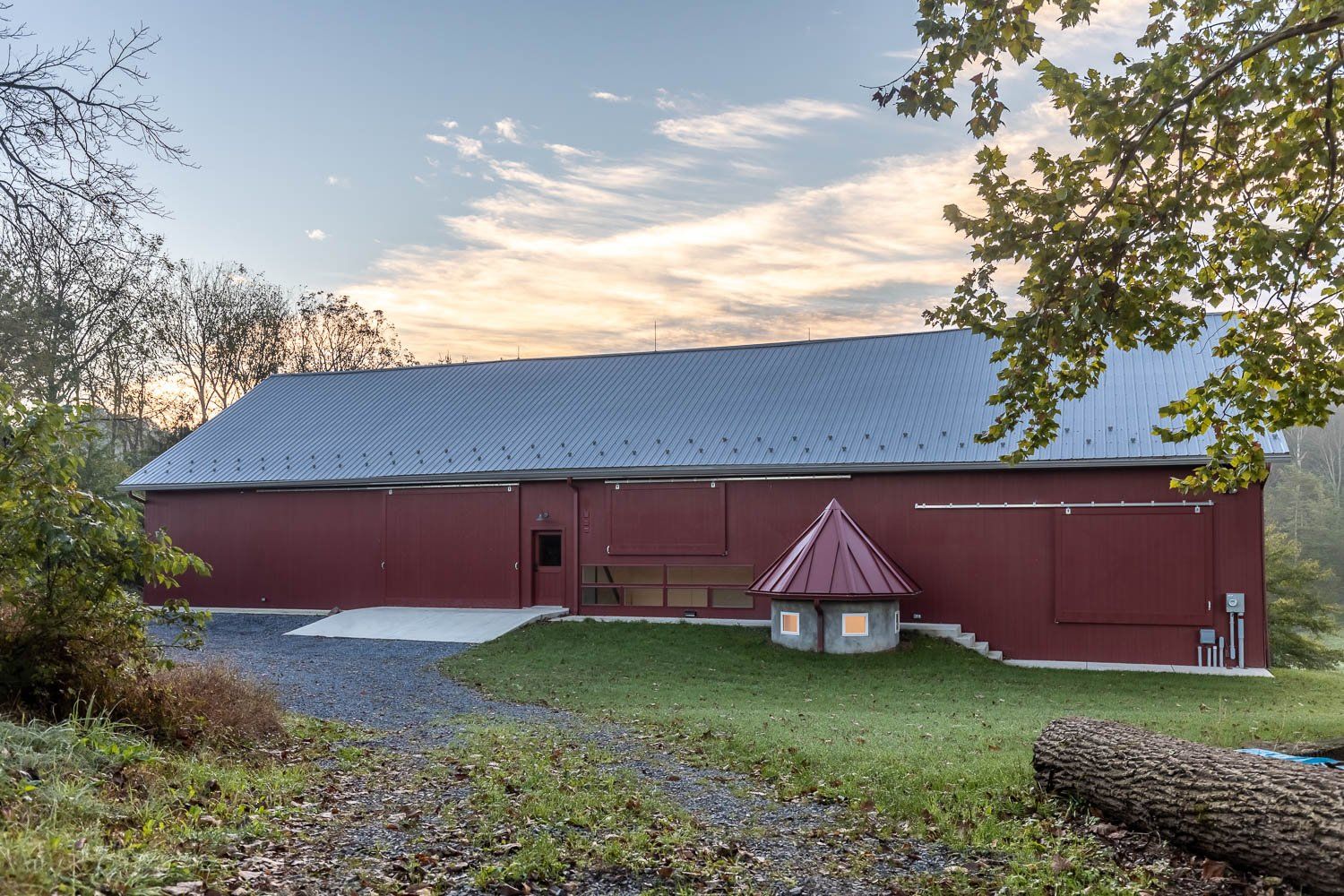 Red barn with a metal roof on a grassy plot. A small, decorative structure sits near the barn.