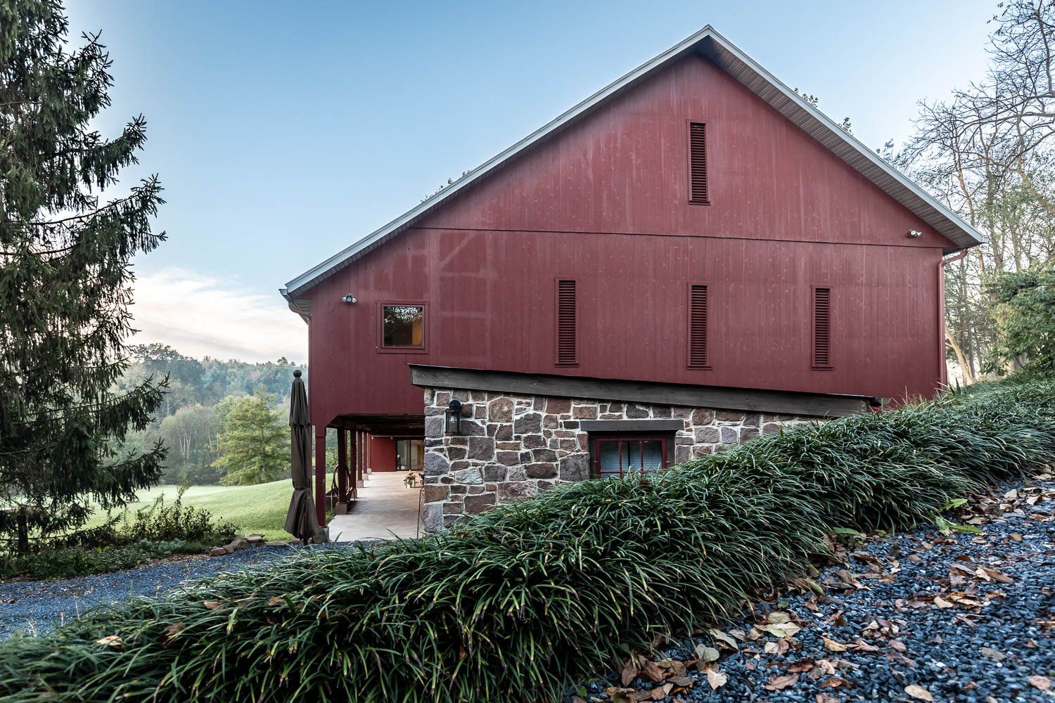 Red barn with stone foundation and greenery in a rural setting under a partly cloudy sky.