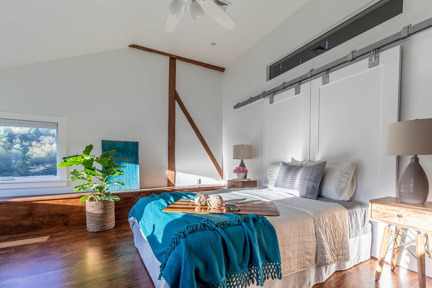 Bright bedroom with bed, wooden floor, and a loft with a plant. White walls, teal throw.