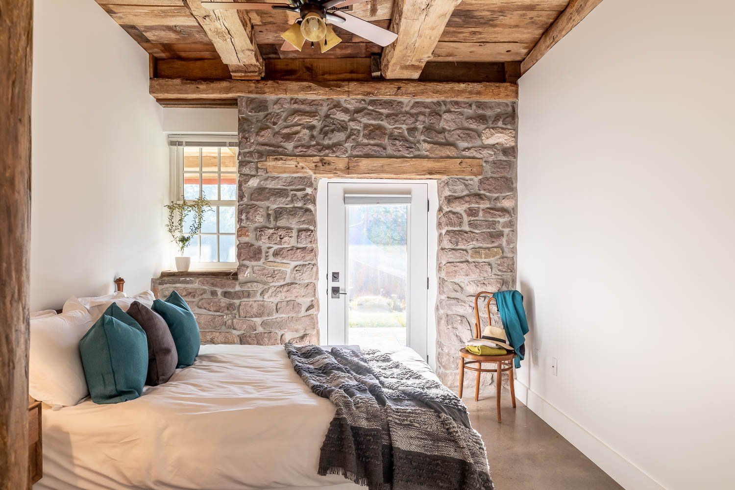 Bedroom with stone wall, exposed wooden beams, bed, open door, and a chair.
