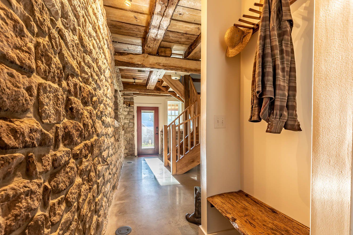 Stone-walled hallway with wood ceiling and staircase. Coat rack with hat and clothes. Sunny interior with view of a door.