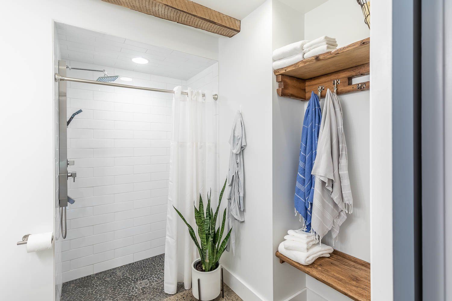Shower with white tiled walls, pebble floor, white curtain, and wooden shelving with towels.