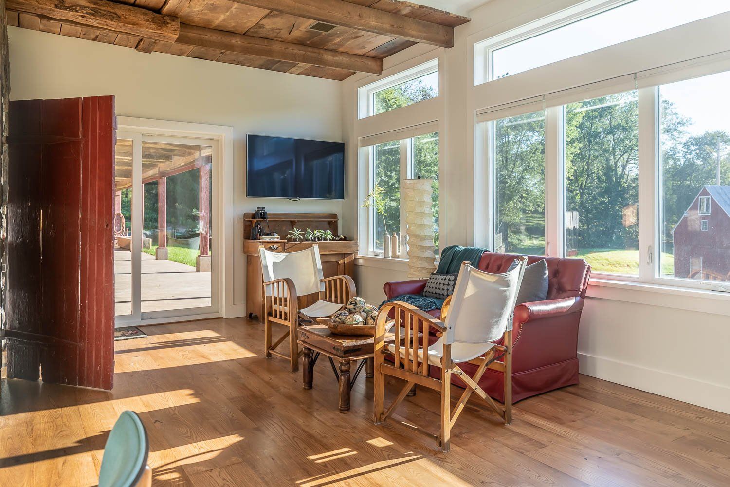 Sunroom with red door, wood beams, seating, large windows overlooking a green yard and a distant red barn.