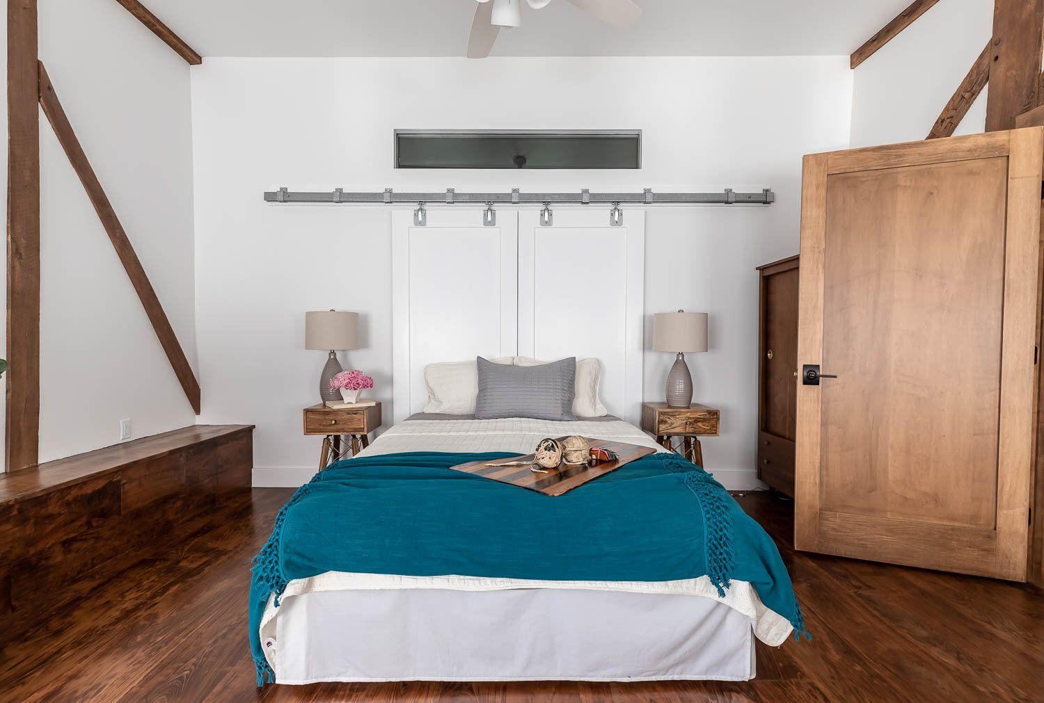 Bedroom with wooden beams, white walls, bed with teal blanket, two bedside tables, open wooden door.