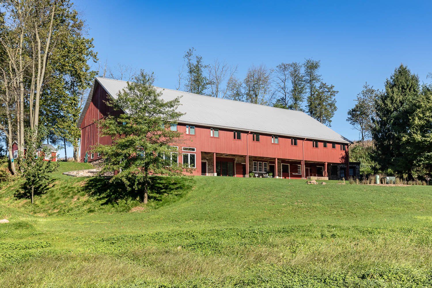 Red barn with a gray roof on a grassy hillside, trees in background, sunny day.