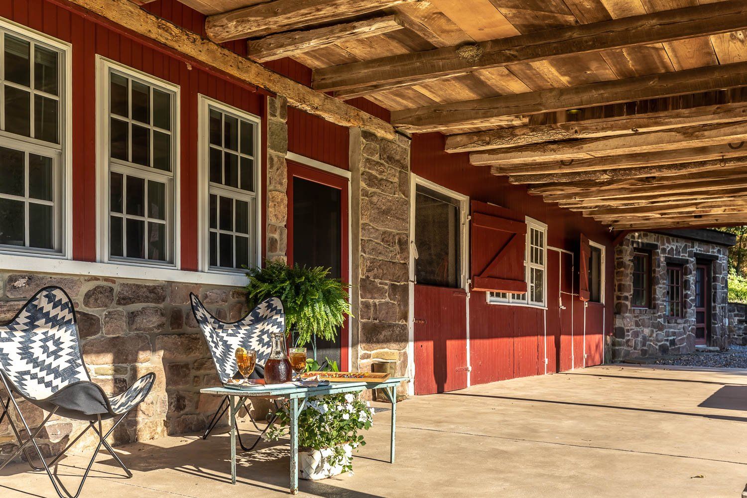 Red barn with stone foundation, wooden beams, windows, patio furniture under a shaded porch.