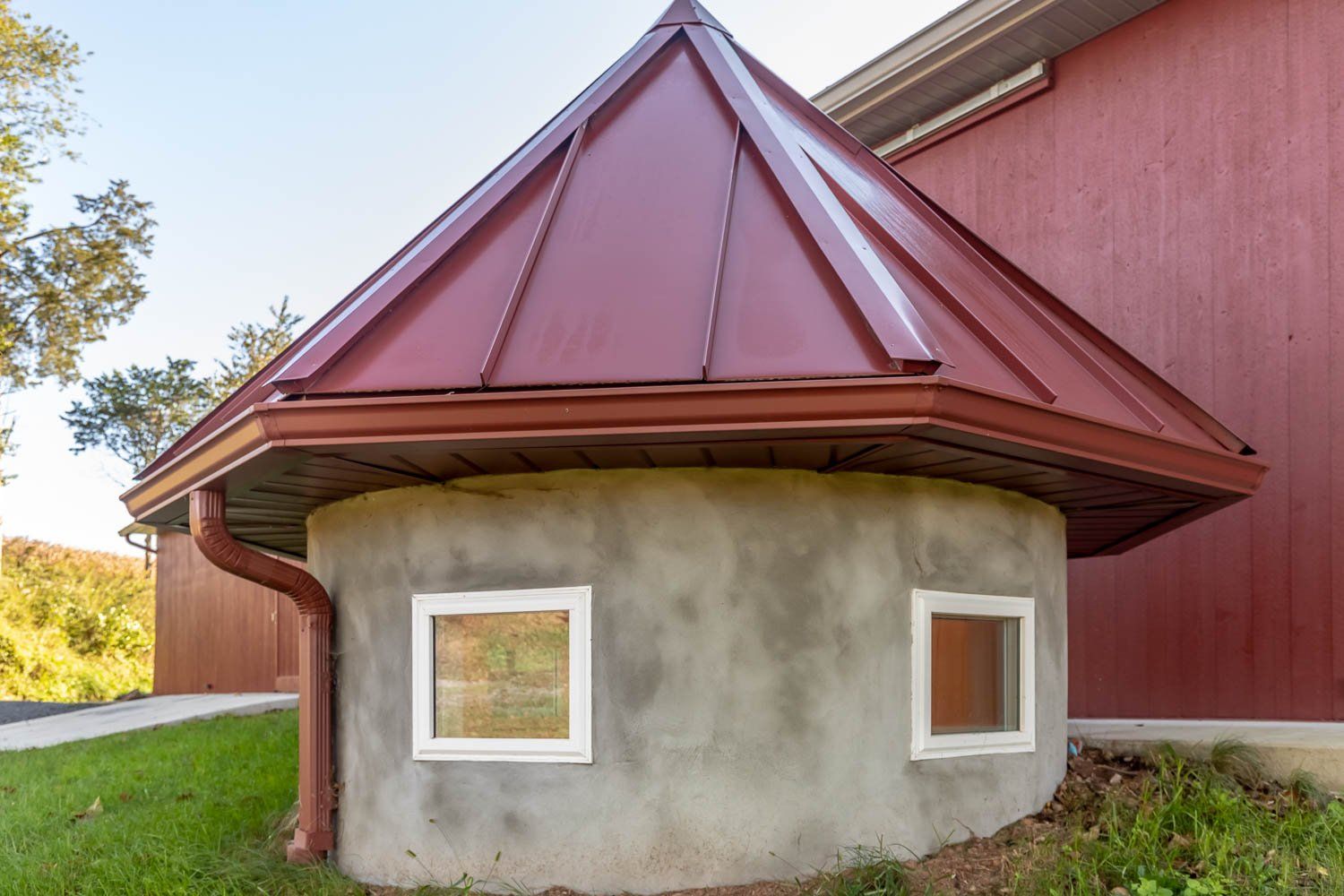 Round, concrete structure with a burgundy metal roof, two square windows, and a red building beside it.
