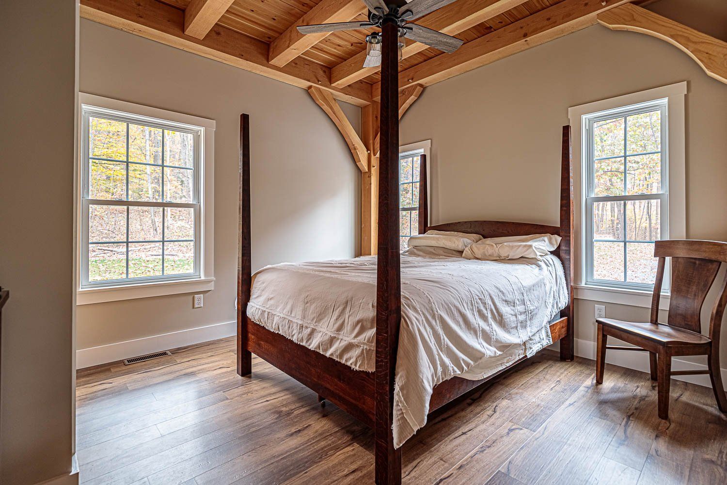 Bedroom with four-poster bed, wood ceiling beams, two windows, and wooden chair on hardwood floor.