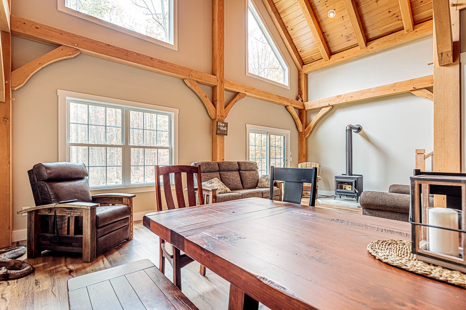 Spacious living room with wood beams, large windows, and a wood-burning stove. Furnished with leather chairs and a dining table.