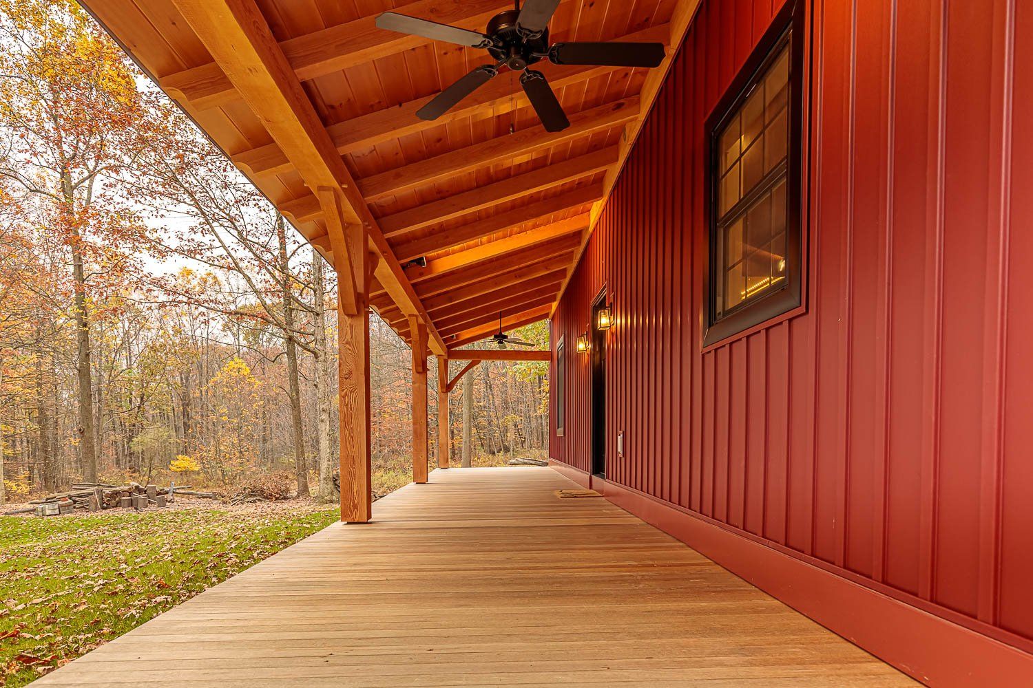 Red-sided house with a wooden porch and a ceiling fan, set in a wooded area with autumn colors.