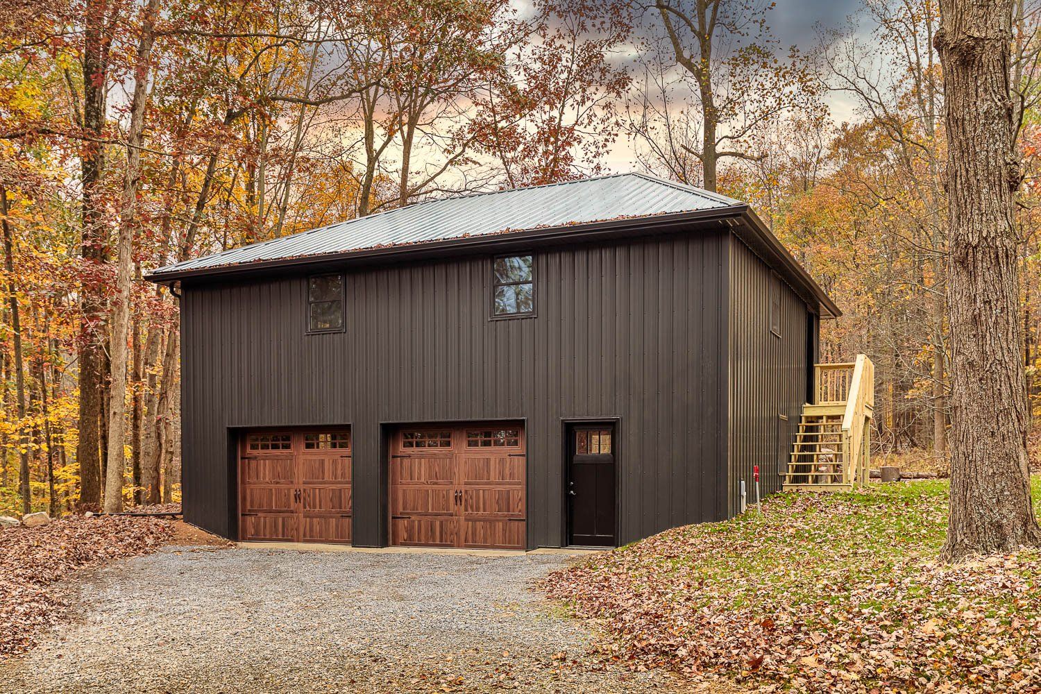 Two-story brown metal building with garage doors and a side staircase in an autumn wooded area.