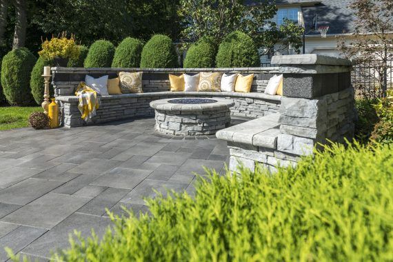 Stone patio with a built-in fire pit and curved seating area, yellow pillows, and green shrubbery.
