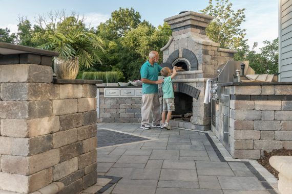 Man and child placing food in a stone pizza oven in an outdoor kitchen.
