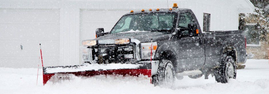 A black truck plows snow in front of a white building on a snowy day.
