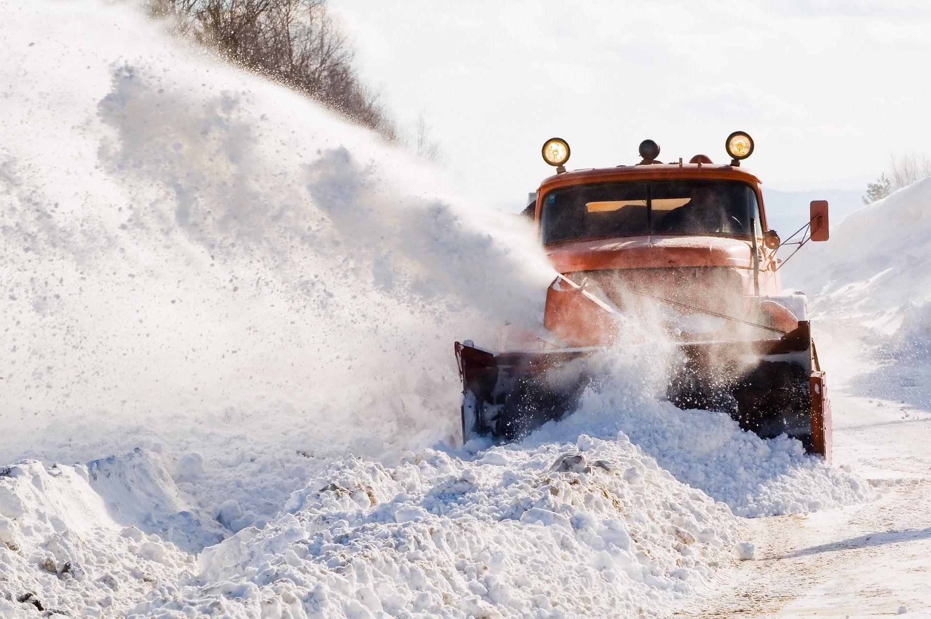 Orange snowplow clearing snow from a road, white snow spraying, bright sunny day.