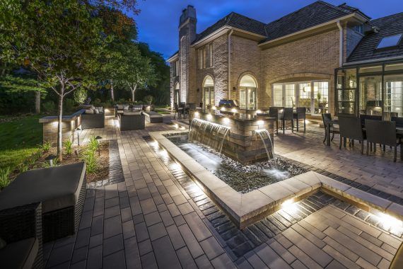 Night view of a luxury backyard patio with water fountain, seating, and a brick house. Lit by warm lights.