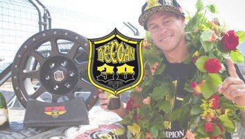 Man wearing floral wreath holds trophy, celebrating a win with a vehicle wheel behind him.