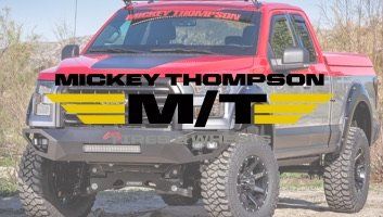 Red and silver Ford pickup truck with Mickey Thompson branding, off-road tires, and modified front bumper.
