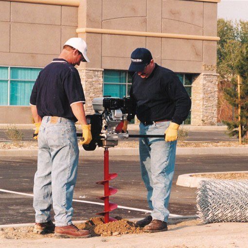 Two workers drilling a hole with a red auger near a building, with a rolled chain-link fence nearby.