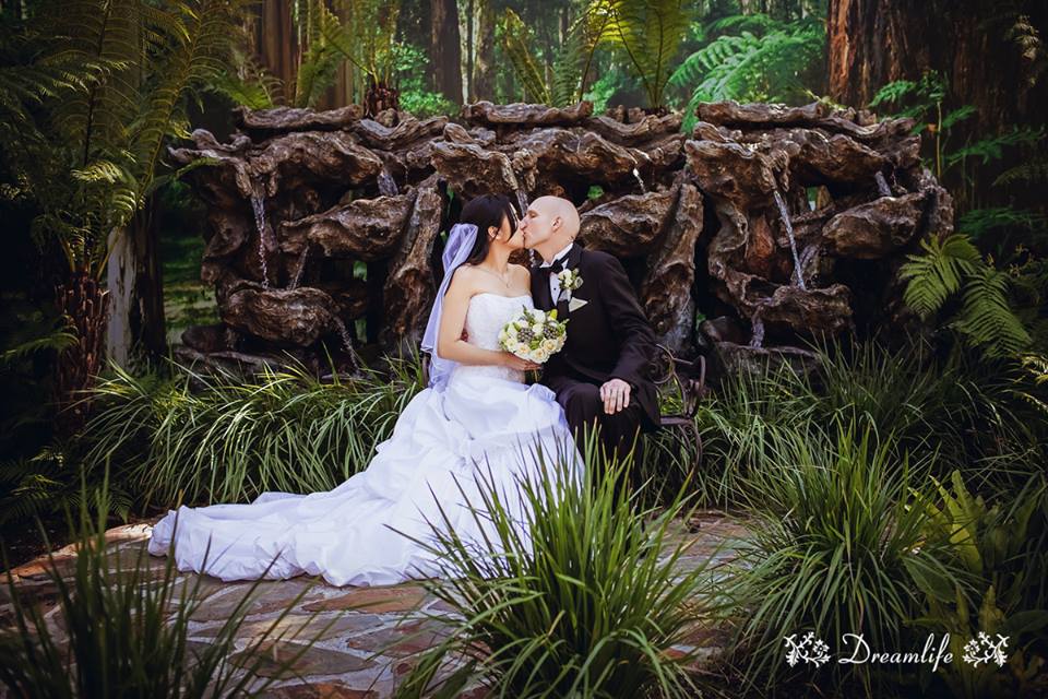 A bride and groom are kissing on a bench in the woods.