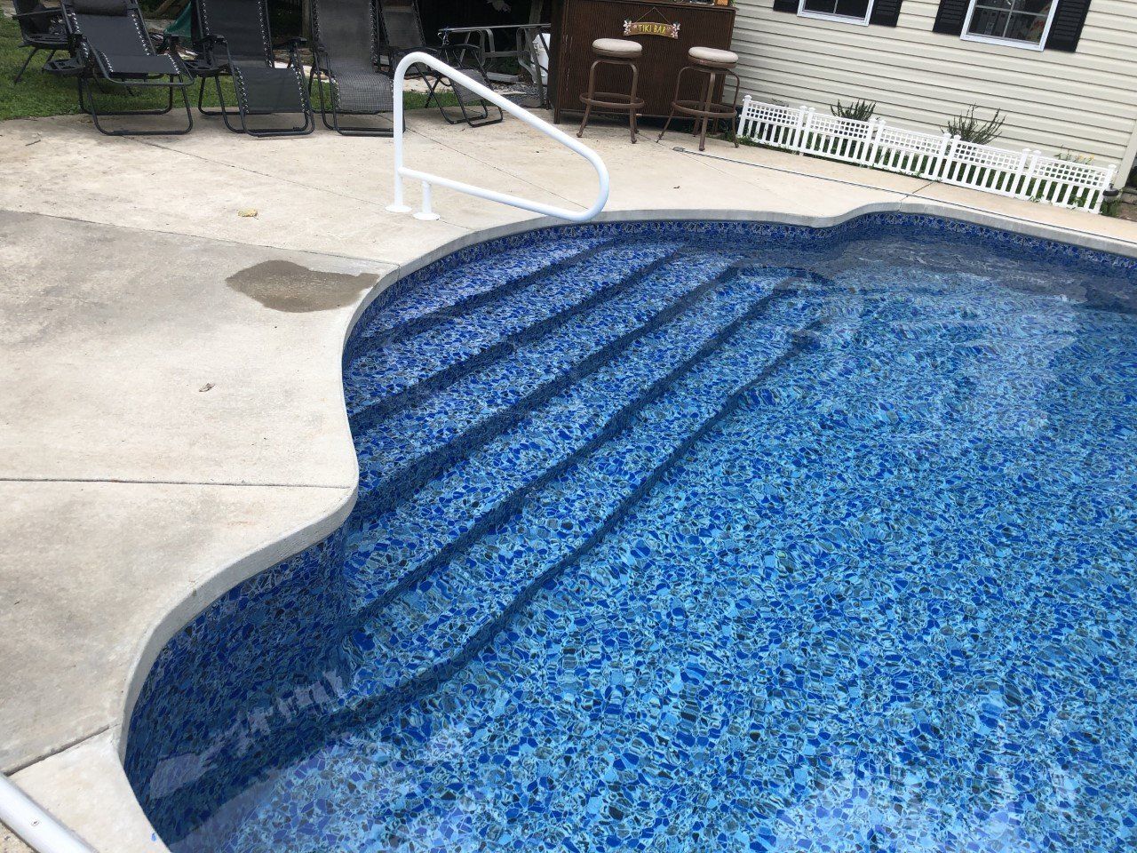 Pool with blue water, steps, and white handrail, set on a concrete patio.