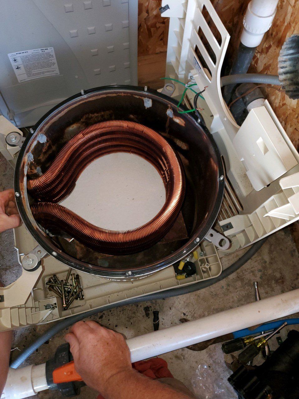 Hands working on a pool heater. Copper coils visible inside the unit. Plastic frame, pipes, and tools are nearby.